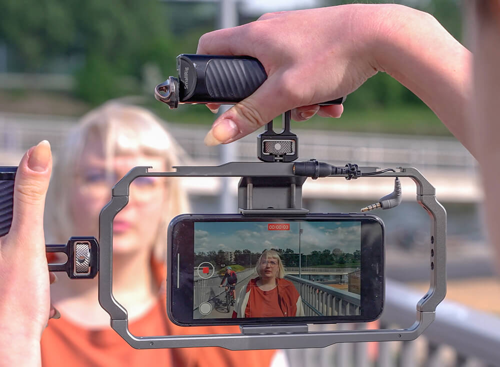 Close-up of a handheld smartphone rig filming a woman on a bridge; an external mic is mounted above the phone.