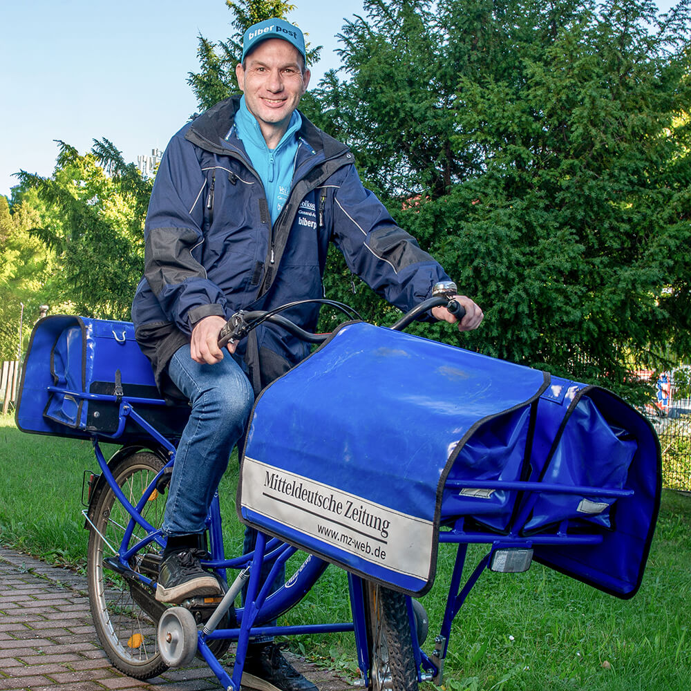 Delivery rider on a blue cargo bicycle delivering newspapers, wearing a blue jacket and cap with Mitteldeutsche Zeitung bag in front park setting.