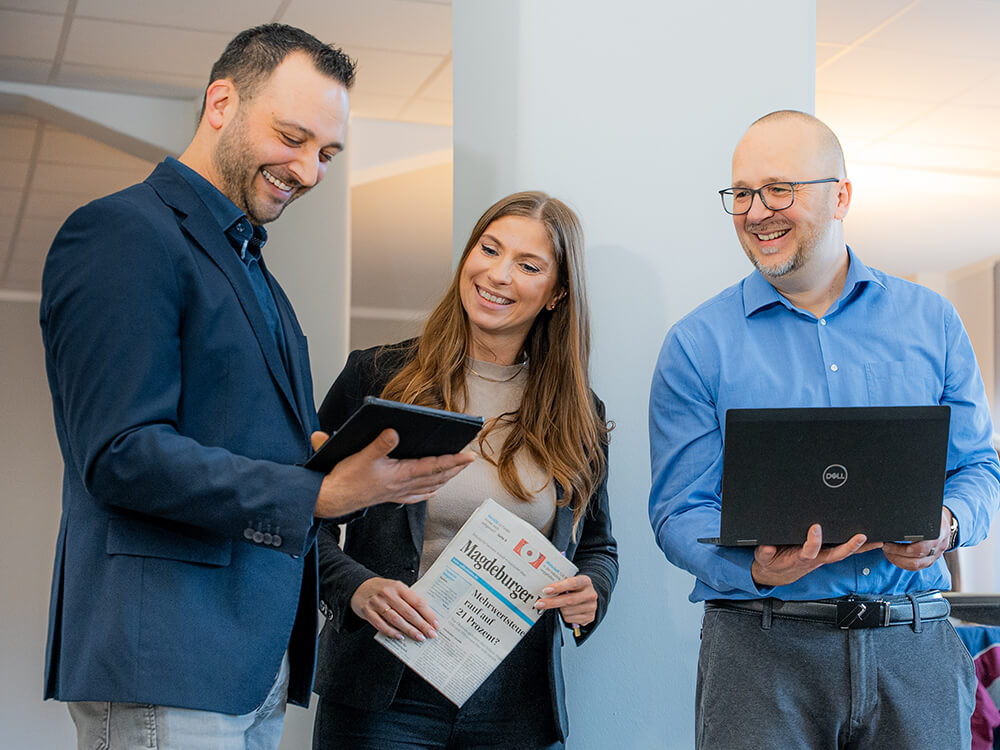 Three coworkers in an office look at a tablet together: woman in the middle holding a newspaper, man on the left in a blazer, man on the right with a Dell laptop.