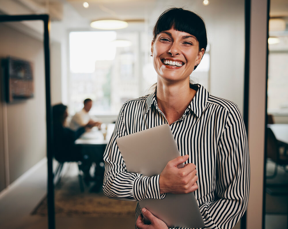 Smiling woman in a striped shirt holding a tablet in a modern office hallway.