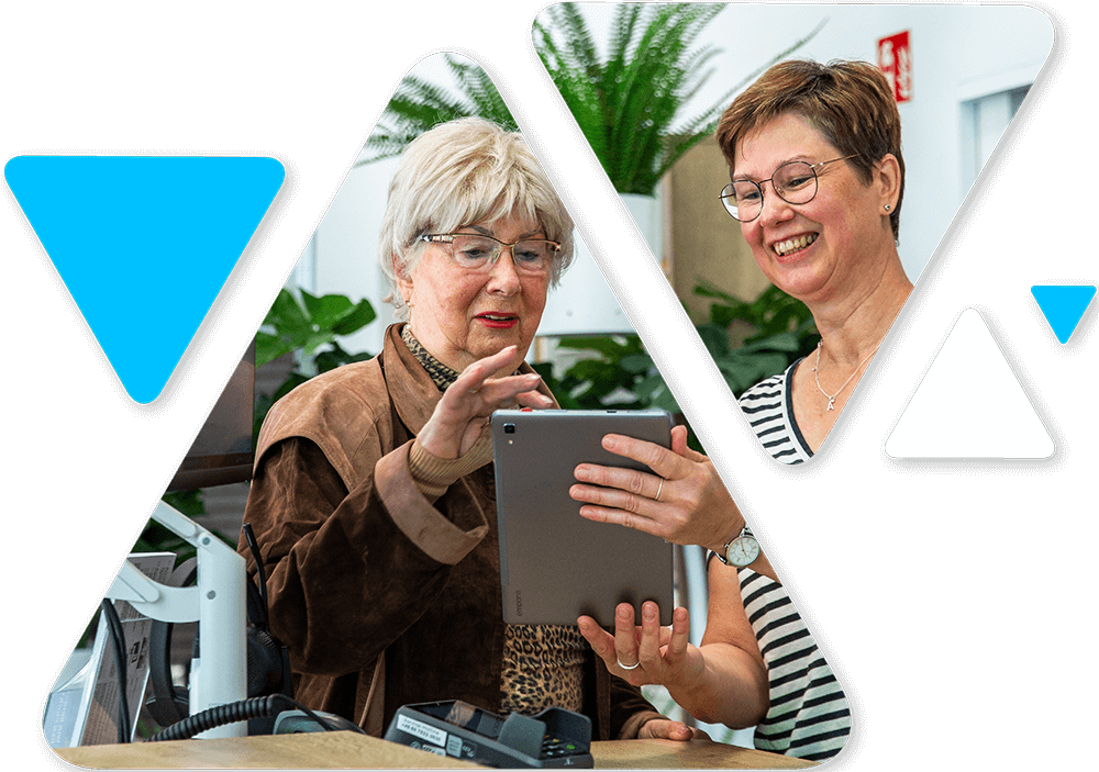 Two smiling women look at a tablet together in a bright office setting with plants in the background.