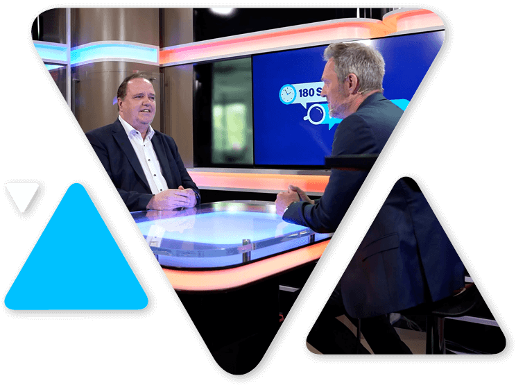 Two men sit at a glass interview desk in a TV studio, talking across from each other with a large screen behind them showing graphics.