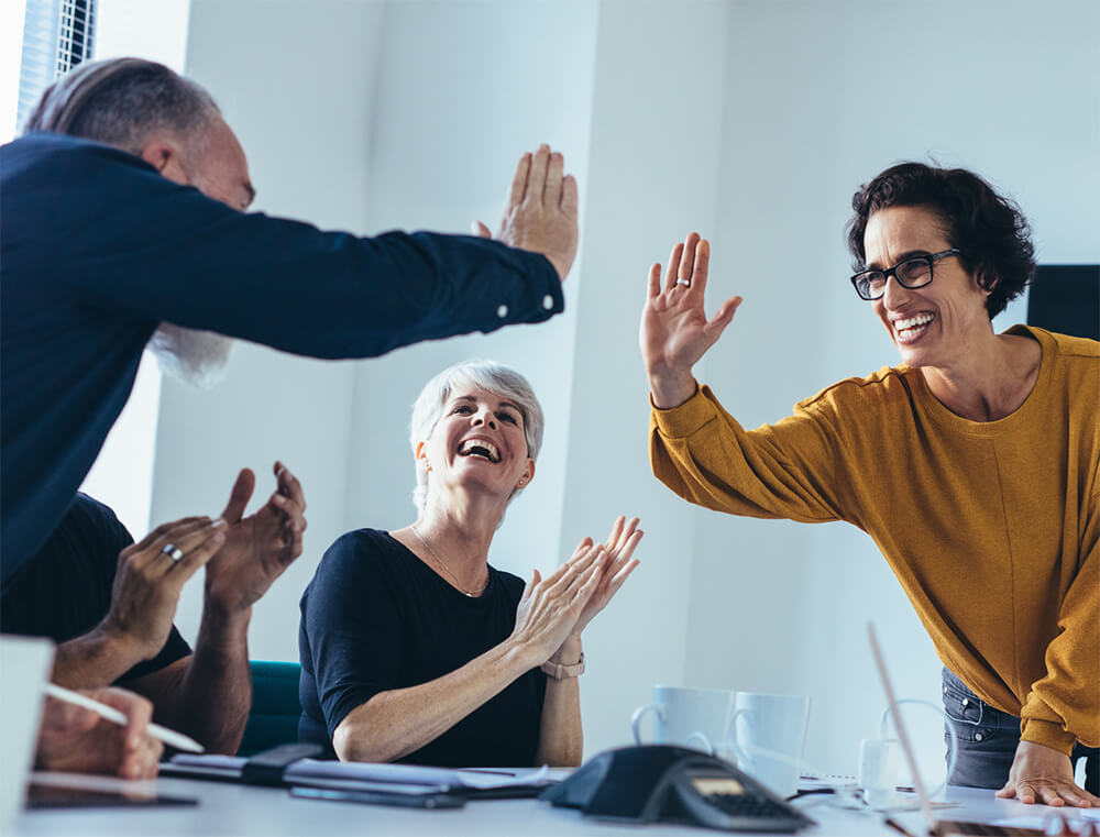 Team of four adults giving a high-five in a bright office around a conference table, celebrating a success.