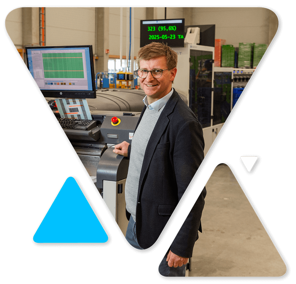 Smiling man in a blazer standing by an industrial control panel in a factory setting.