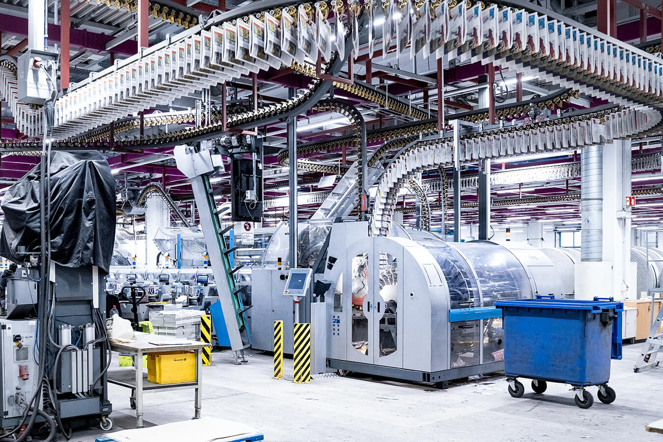 Industrial factory floor with large automated production line and overhead conveyor systems in a bright, busy facility.