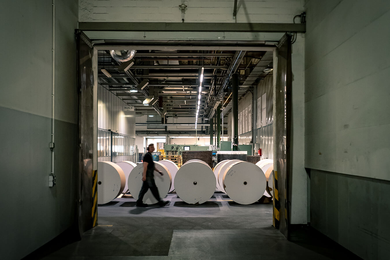 Industrial warehouse with large white paper rolls lined up; a worker walks past them toward the back.