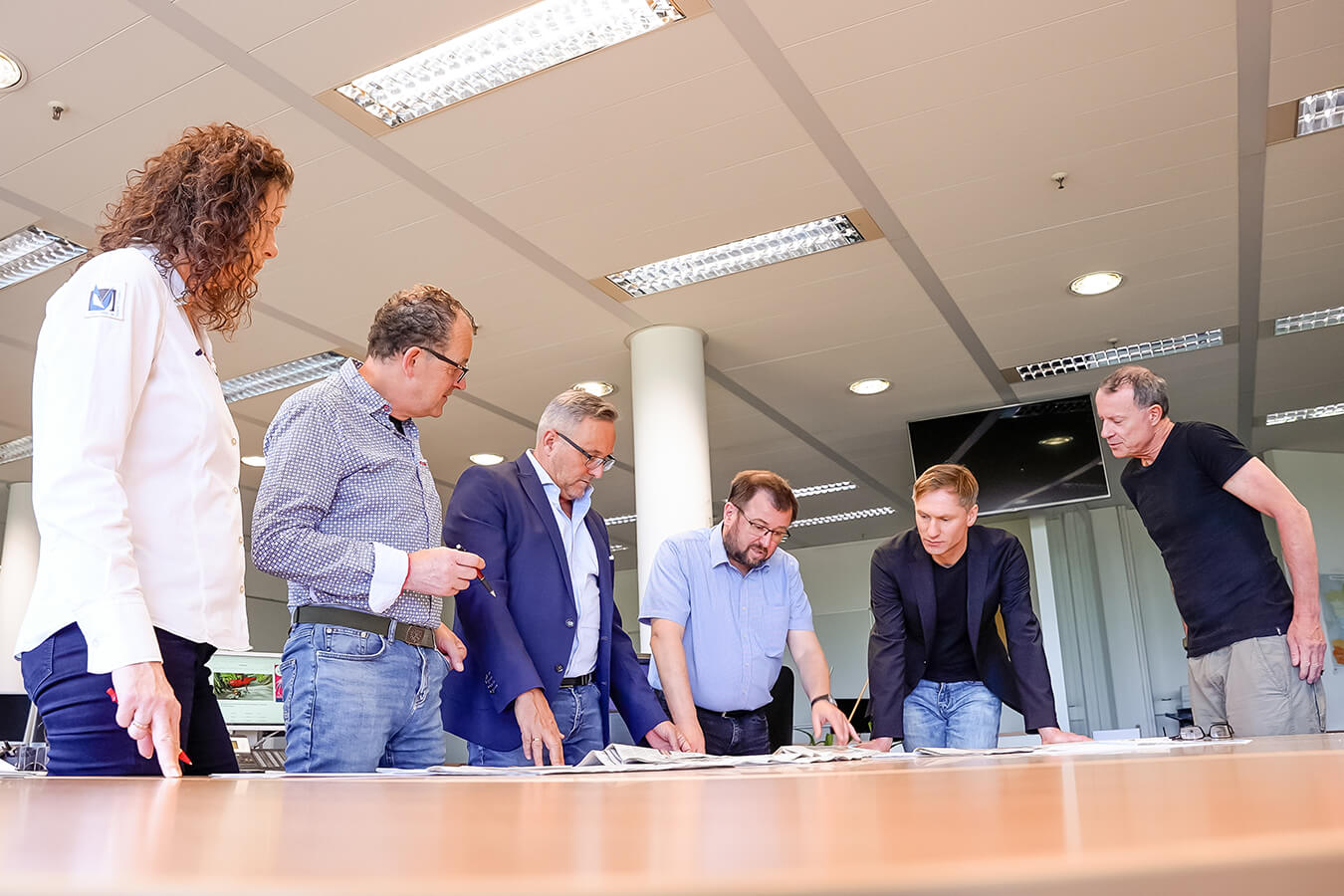 Six colleagues (one woman and five men) stand around a conference table, examining papers laid out before them in a bright office.