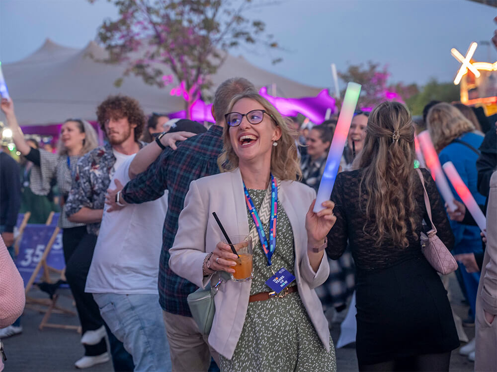 Smiling woman in glasses and a beige blazer holding an orange drink with a straw at a crowded outdoor festival, waving a glow stick.