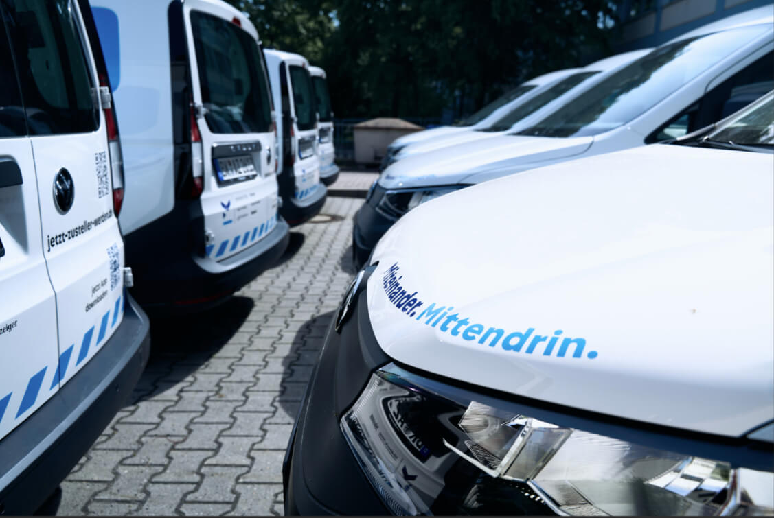Fleet of white vans with blue branding parked in a parking lot, foreground showing the hood with 'Mit ten drin' text.