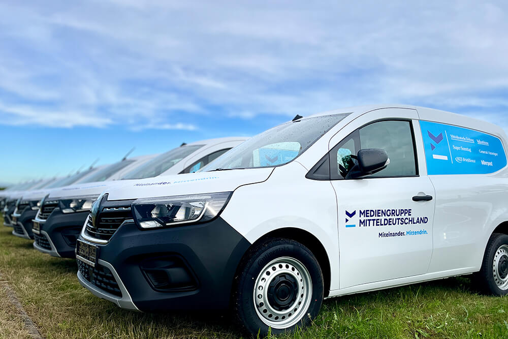 Row of white delivery vans with blue logos parked on a grassy field under a blue sky, branding reads Mediengruppe Mitteldeutschland.