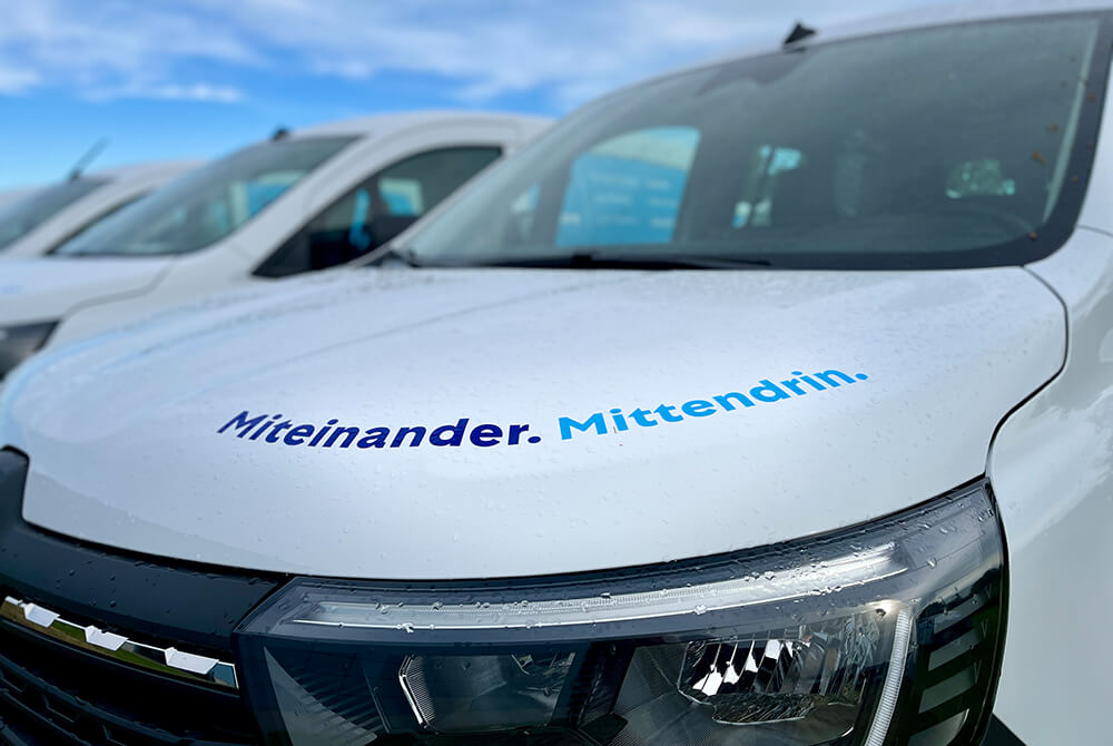 White van with blue German slogan 'Miteinander. Mitten drin.' on the hood, parked with other vans nearby on a sunny day.